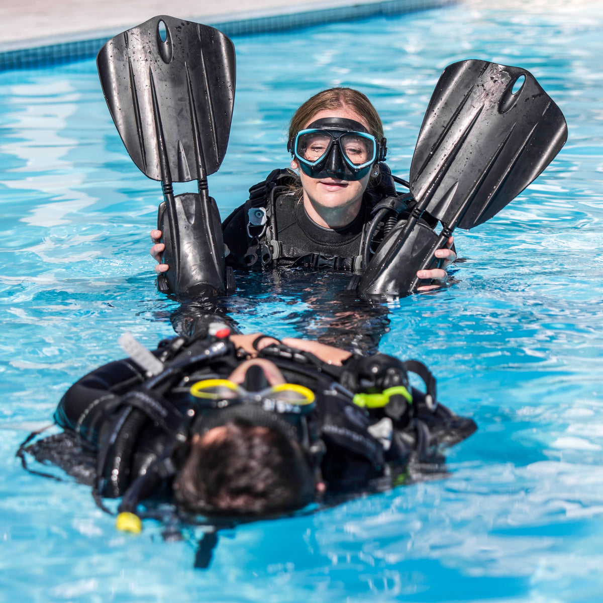 A person in diving gear is being assisted at the surface by an instructor who is holding their fins and mask.